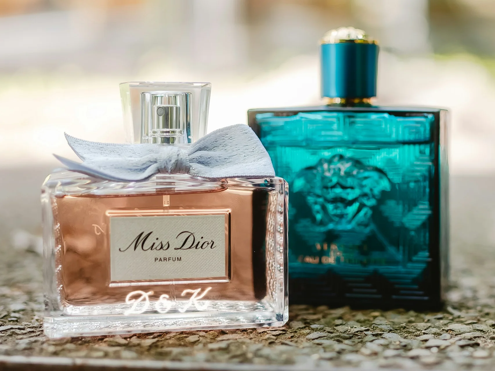 Miss Dior bow-capped bottle beside teal Versace Eros bottle on stone surface, side-by-side longevity comparison still life